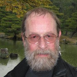 A bearded and bespectacled man, Professor David Ormandy, poses in front of a forest and lake.